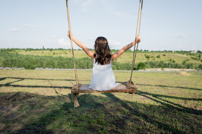 back view of child in summer dress riding swing in meadow